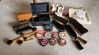 Full overview of shoe shining supplies including brushes, polish tins, cloths, and cases laid out on carpeted floor