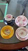 Four assorted tea cups and saucers with floral patterns and gold accents on a glass table.