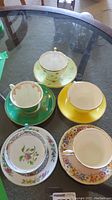 Five tea cups each with matching saucers in various floral and gold-trimmed patterns placed on a glass table.