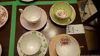 Four teacups and saucers shown from above on a table with identification tag. Each cup and saucer has a different design including shamrocks, floral, and pink solid colors with gold trim.