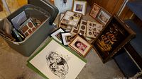 Wide shot showing art prints arranged on the floor around a large grey plastic bin containing picture frames and other prints. Includes the velvet ship painting leaning against a wall and multiple frame sets scattered nearby.