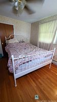White metal bed frame shown assembled in a bedroom with patterned bedding. Shows headboard, footboard, and side rails.