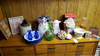 Wide view of dresser top with all displayed items including Capodimonte vase, whiskey jug, cobalt vases, floral ceramics, framed prints.