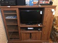 Front view of the entertainment cabinet showing the wood grain laminate finish, glass door with shelves on left side, central open shelves with TV, and enclosed cabinet on right.