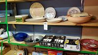 Wide view showing three boxes of flatware, collector plates, pink ceramic bowl, and other kitchenware on shelves