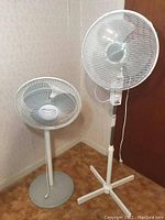 Two floor fans placed in corner, one shorter with grey pedestal base, one taller with white four-legged base.