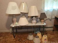 Six lamps with bases displayed on a white folding table in front of a window and wall with a heating element. Lamps vary in style and height ranging from about 14" to 24".