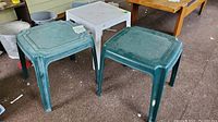 Three plastic patio side tables grouped together - two green and one white, showing tops and legs with staining and wear.