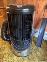 Front and side view of black and stainless steel cylindrical portable air conditioner with vent and grille visible, standing on a carpeted floor against panelled wall.