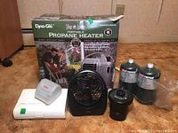 Grouping of all lot items showing the propane heater box, two propane bottles, blood pressure monitors, O2 Cool fan, and Intex Quick-Fill device on wood floor against plain wall.