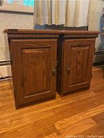 Pair of wooden side table cabinets shown side by side on wooden floor with curtains in background. Both tables have paneled doors with ornate metal handles and carved detailing on top edges.