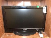 Full front view of Dynex LCD TV including base stand and remote control placed on the surface next to it. Shows overall condition and size.