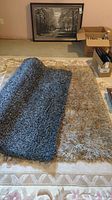 Beige and grey shag area rugs positioned on a patterned floor rug, showing texture and size comparison, beige rug is smaller and grey rug is larger and folded at one corner