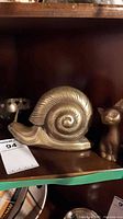 Brass snail figurine with detailed spiral shell and part of cat figurine beside it on shelf