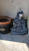 Photo showing sitting Buddha statue with noticeable paint chipping on head and face, seated on ground next to two garden pots