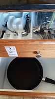 Open drawer showing white plastic cutlery tray holding assorted stainless steel forks, spoons, and knives, next to a black frying pan with plastic handle beneath the drawer.