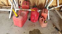 Five red plastic gas cans in varying sizes and shapes, placed on concrete floor against plywood wall.