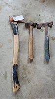 Photo of an axe with a damaged wooden handle and two hammers laid on a concrete floor.
