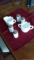 Full set of hobnail milk glass items including one cream pitcher, one sugar bowl, two salt shakers, and two pepper shakers arranged on a red cloth