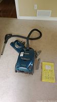 Blue Kenmore Elegance canister vacuum on beige carpet with hose, wand, and additional attachments and instruction manual nearby