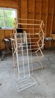 Wooden foldable drying rack with two white metal wire shelves leaning against it in a garage or workshop environment.