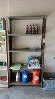 Full view of metal shelving unit showing four shelves with various items including cleaning sprays, helmet, watering cans, and other small items.