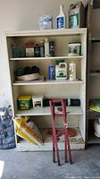 Full view of white metal garage shelf containing garden and lawn care products with large white bag on bottom shelf, hand tools in front, and multiple small bottles and containers spread across three shelves.