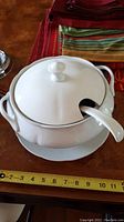 White ceramic soup terrine covered with matching lid and ceramic ladle resting inside. Mounted on a round ceramic plate. Approximate diameter 10 inches shown by ruler.