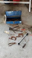 Blue metal toolbox and assorted hand tools including clamps, screwdrivers, pliers, and ratchet parts spread on floor