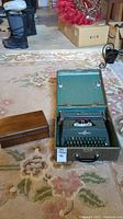Green Underwood typewriter inside matching green carrying case placed on floral carpet alongside polished vintage wooden box. Background shows boots and boxes.