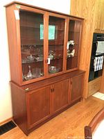 Full view of the two-piece teak wood china cabinet placed against a wall in a kitchen or dining area, showing upper glass display and lower wooden storage units.