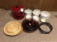 Full set of cookware displayed on carpeted floor showing the large red pot, three white ceramic cooking dishes with lids, stacked dark glass bowls, woven basket liners, and two small white oval dishes. Included a garlic bulb for scale.