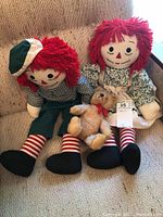 Photo of two Raggedy Ann and Andy dolls sitting on a beige couch with a small brown teddy bear with red bow placed in front between them.