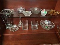 Display of various glass and crystal bowls, candy dishes, bell, and other decorative glass items arranged on shelves.
