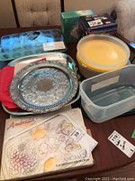 Overview of lot showing silver serving platters, yellow mixing bowls, clear food storage container, and kitchen towels with fruit designs