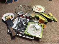 Wide shot of lot showing cleaver, cutlery, collector plates, ceramic corn servers, mugs, and utensils on carpet.
