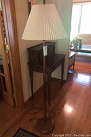 Full view of wooden floor lamp with round base and pleated paper shade, next to wooden furniture and door frame.