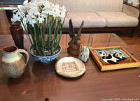 Photo of the entire lot displayed on a glass table: pottery jug, carved wooden soldier, carved stone owl, pottery ashtray, faux white flowers in porcelain bowl, and framed yarn craft panda art.