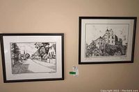 Two framed prints hanging on the wall: one showing a street scene with houses and a person walking, the print is signed and numbered 90/195 in the lower left corner. The second print shows a large architectural building with trees, signed on the lower right.