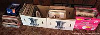 Three cardboard boxes and loose stacks filled with vinyl records on carpeted floor