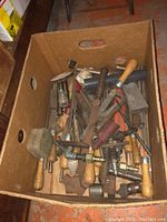 Box filled with assorted hand tools, mainly with wooden handles, including files, chisels and a mallet, showing wear and possible rust.