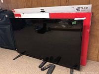 48 inch RCA LED TV standing on carpet with three remote controls in front, original packaging behind.