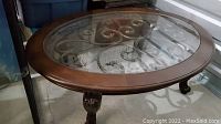 Oval wood framed coffee table with removable glass top showing ornate faux wrought iron design beneath glass, sitting on carved claw feet legs.