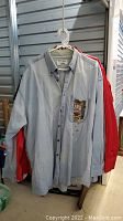 Front view of a light blue denim long-sleeve button-up shirt with embroidery showing a slot machine on the chest pocket. Red and white T-shirts are partially visible behind it.