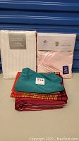 Photo showing white damask stripe tablecloth and napkins package, pink pillowcases package, and assorted folded linens including bright teal and red striped patterns.