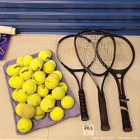 Three tennis racquets and multiple tennis balls placed on a surface with a blue background. Racquets arranged side by side with wear visible on handles.