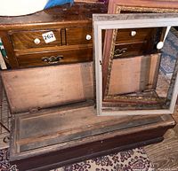 Antique wooden tool chest with open lid revealing gray interior, three assorted wood frames leaning on it, and wooden three-drawer chest behind.
