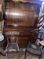 Photo showing solid wood cabinet with multiple drawers and cupboard doors, with two vintage chairs placed in front of it, one with floral seat and curved backrest, the other with spindle backrest.