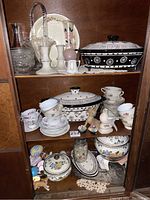 Three shelves of cabinet contents showing ceramic casseroles, teacups, figurines, glassware and decorative items.
