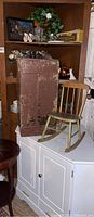 Brown wood corner cabinet with shelves and closed storage, brown chest in front, and green child-sized rocker on top, along with white corner cabinet below.